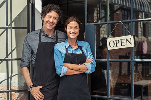 Smiling business owners in front of store next to open sign and wearing protective aprons.